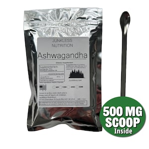 Op de afbeelding: Een zilveren zakje met Ashwagandha voedingssupplement van Junkless Nutrition, met een 500 mg schep. Het etiket bevat voedingsfeiten en gebruiksaanwijzingen. Naast het zakje ligt een lange, zilveren lepel.