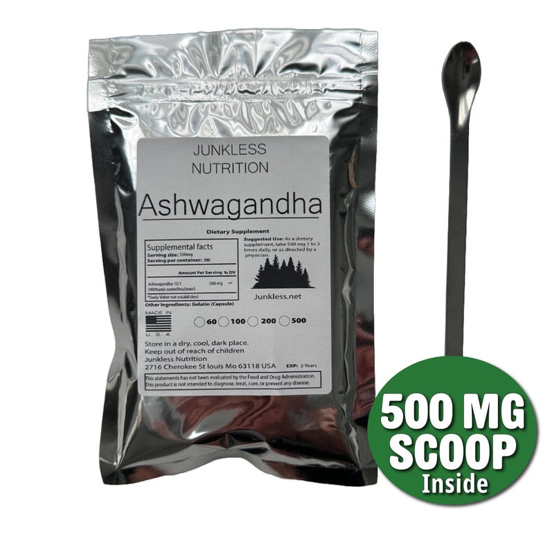 Op de afbeelding: Een zilveren zakje met Ashwagandha voedingssupplement van Junkless Nutrition, met een 500 mg schep. Het etiket bevat voedingsfeiten en gebruiksaanwijzingen. Naast het zakje ligt een lange, zilveren lepel.