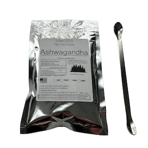 Op de afbeelding: Een zilveren foliezakje met Ashwagandha voedingssupplement, een wit etiket en een metalen lepel. Het etiket bevat de merknaam "Junkless Nutrition". De lepel is lang en slank.