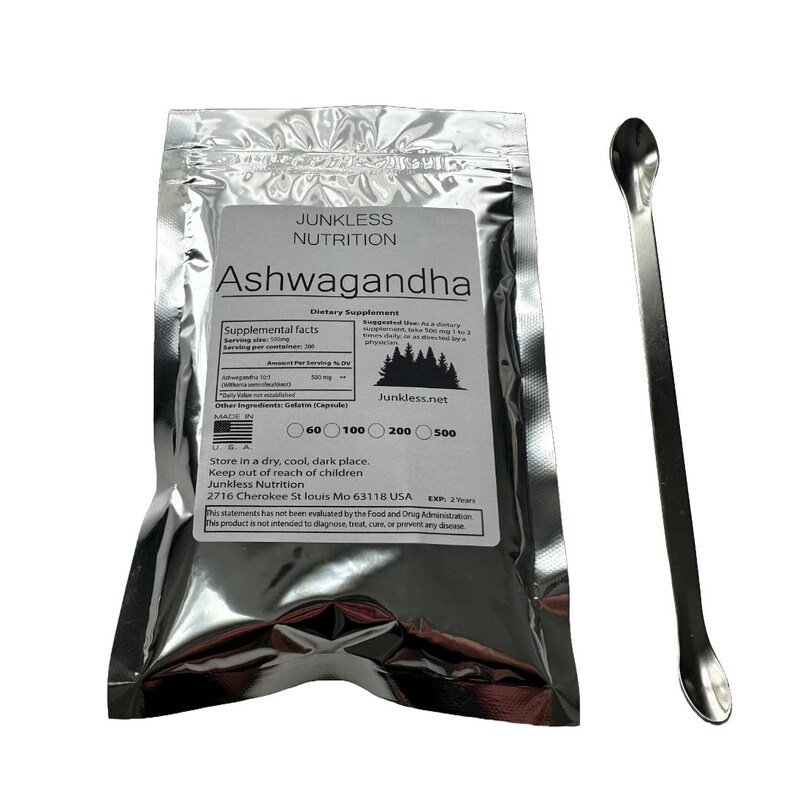 Op de afbeelding: Een zilveren foliezakje met Ashwagandha voedingssupplement, een wit etiket en een metalen lepel. Het etiket bevat de merknaam "Junkless Nutrition". De lepel is lang en slank.