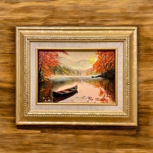 May include: Oil painting of a lake scene with a small boat, framed in a gold-colored frame. The artwork depicts a serene autumn landscape with vibrant red and orange trees reflected in the water. The painting is displayed on a wooden surface.
