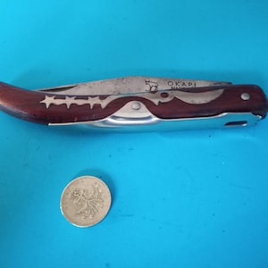 May include: A vintage folding knife with a dark wood handle and a stainless steel blade. The handle features a row of white stars. The blade is marked "OKAPI". A silver coin is next to the knife.