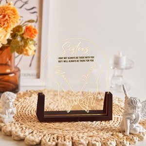 May include: A wooden base with a clear acrylic panel featuring a gold outline of two hands holding each other. The text "Sisters" is at the top of the panel, followed by the text "I may not always be there with you but I will always be there for you." The names "Aylmer" and "Darris" are written below the hands.