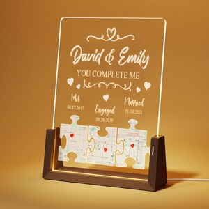 May include: A white acrylic plaque with a wooden base, featuring the names "David & Emily" and the text "You Complete Me". The plaque is decorated with hearts and a puzzle piece design, with dates for "Met", "Engaged", and "Married".