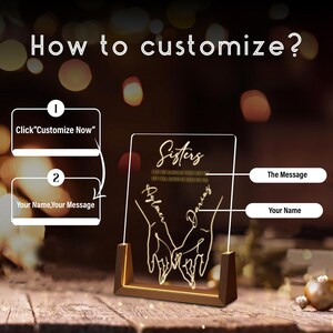 May include: A personalised illuminated acrylic sign with a wooden base. The sign features a design of two hands holding each other with the word "Sisters" and a message about always being there for each other. The sign can be customised with names and a message.