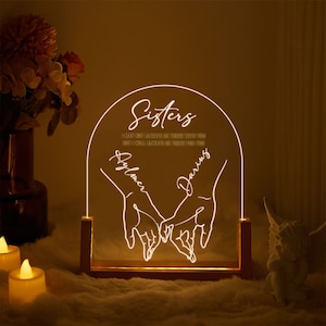 May include: A white LED light with a wooden base features a design of two hands holding each other with the text "Sisters" and the quote "I may not always be there with you but I will always be there for you".
