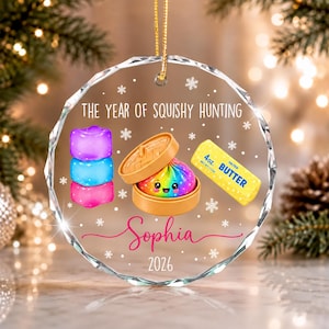 May include: A circular glass ornament featuring the text THE YEAR OF SQUISHY HUNTING, Sophia, and 2026. The design includes a stack of purple and blue cubes, a rainbow-colored dumpling in a bamboo steamer, and a yellow 4oz. salted butter stick.