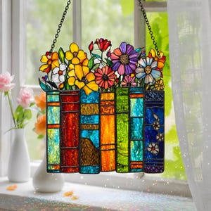 Könnte beinhalten: Buntes Glasfenster-Hängebild mit einer Reihe von Büchern und bunten Blumen. Die Bücher sind in Rot, Blau, Orange und Grün gehalten. Die Blumen sind gelb, weiß, rot und lila. Das Fenster wird von einer schwarzen Kette gehalten.