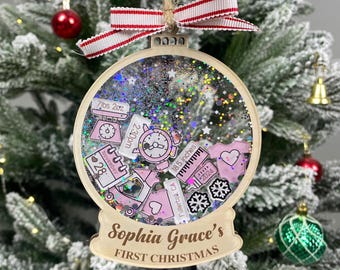 May include: A round wooden Christmas ornament filled with holographic glitter and various pink and white baby-themed charms, including a scale labeled 7lbs 2oz, a clock, a calendar showing November 28, and a bottle. It reads Sophia Grace's FIRST CHRISTMAS.