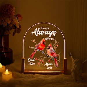 May include: A wooden illuminated sign with a white arch and the text "We are Always with you". Two red cardinals are perched on a branch with red berries. The sign also includes the text "Dad 2020" and "Mom 2021".