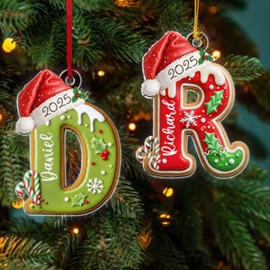May include: Two Christmas ornaments shaped like gingerbread letters, "D" and "R", are displayed against a backdrop of a Christmas tree. The ornaments are decorated with red and green icing, holly, and candy canes. Each ornament has a Santa hat with the year "2025" and a name.