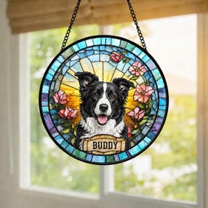 May include: Round stained glass window art featuring a black and white Border Collie dog with the name "Buddy" on a banner. The design includes pink flowers and a colorful mosaic border, hanging from a black chain.