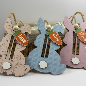 May include: Three wooden bunny-shaped ornaments with a carrot design and personalized names. The bunnies are pink, blue, and light brown with floral and geometric patterns. The names are "Luna", "Nacy", and "Sydney".