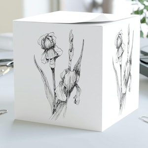 May include: A white cube-shaped notepad with a black and white line drawing of iris flowers on the front.