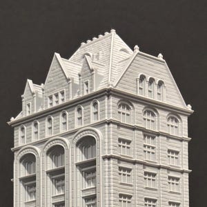 Op de afbeelding: Een gedetailleerd, lichtgrijs miniatuurmodel van een hoog gebouw met meerdere verdiepingen, ramen en architectonische details. Het model heeft een puntdak en boogvensters, wat een klassiek ontwerp laat zien. Het gebouw staat tegen een donkere achtergrond.
