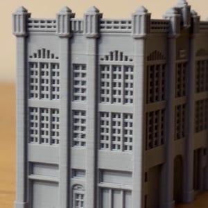 Può includere: Un modello di edificio in miniatura grigio stampato in 3D caratterizzato da una facciata a più piani con colonne verticali, numerose finestre rettangolari, un tetto piatto con parapetto decorativo e porte al piano terra.