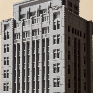 May include: A gray, three-dimensional scale model of a tall skyscraper with a rectangular footprint. The structure features a grid of recessed windows, vertical columns, and a stepped roof design, all rendered in a solid, matte gray finish.