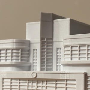 May include: A white 3D printed architectural model of a building featuring a central tower section flanked by two symmetrical wings with grid-patterned windows and a low-profile front entrance section with a small circular detail above the door.