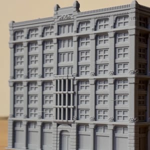 Puede incluir: Un modelo gris impreso en 3D de un edificio comercial de varias plantas que presenta una fachada simétrica con patrones de ventanas repetitivos, columnas verticales, cornisas decorativas y una sección de entrada central con una puerta arqueada.