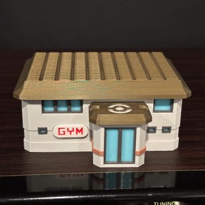 May include: A 3D-printed model of a building with a tan roof and white walls. The building has blue windows and a sign that says "GYM" in red. The entrance features a symbol and blue doors.