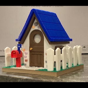 Peut inclure: Une petite maison blanche avec un toit bleu, une porte brune et une boîte aux lettres rouge. La maison a une clôture en bois blanc et repose sur une base verte.