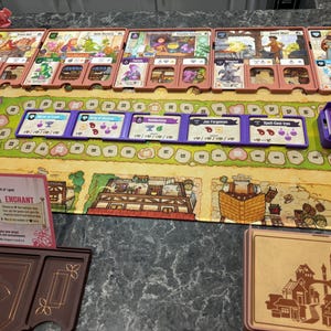 May include: A board game setup with various cards, tokens, and a game board. The cards are in pink holders, and the board features a green path with circular markers. Purple trays and tokens are also visible, suggesting a strategy game.