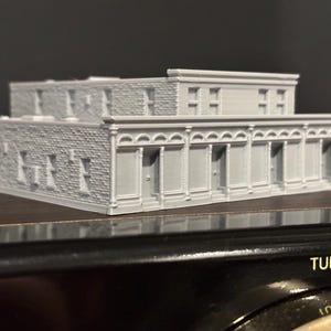 Op de afbeelding: Een grijs, 3D-geprint model van een gebouw met twee verdiepingen en een bakstenen gevel. Het gebouw heeft meerdere ramen en deuren, met decoratieve afwerking langs de daklijn en boven de deuren. Het model staat op een donkere ondergrond.