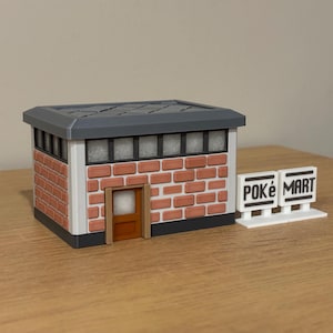 May include: A miniature model of a brick building with a gray roof, a brown door, and a row of windows. Next to the building are two white rectangular signs with black text that read POKÉ MART, featuring a black border and a white base stand.