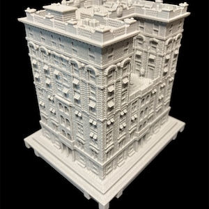K&ouml;nnte beinhalten: Ein wei&szlig;es 3D-gedrucktes Modell eines hohen, schmalen Geb&auml;udes mit vielen Fenstern und komplizierten Details. Das Geb&auml;ude hat einen quadratischen Sockel und ein flaches Dach.