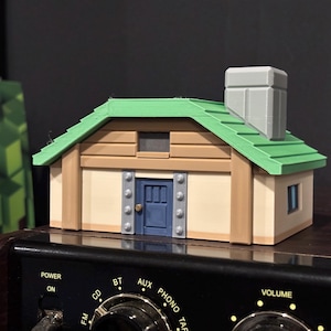 May include: A miniature house model with a green roof, tan walls, and a blue door. The model has a gray chimney and is sitting on top of a stereo system. The house has a small window and is made of plastic.