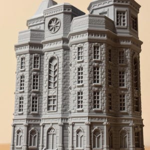 May include: A grey, three-dimensional printed model of a multi-storey building featuring a brick-textured facade, numerous rectangular windows, arched ground-floor entrances, a central circular clock-like detail, and two prominent roof towers.