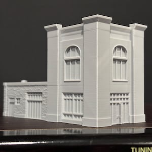 Op de afbeelding: Een lichtgrijs architectonisch model van een gebouw met boog- en rechthoekige ramen. Het model heeft een gedetailleerde gevel, inclusief een bakstenen gedeelte en een hoofdstructuur met een plat dak. Het is een miniatuurreplica.