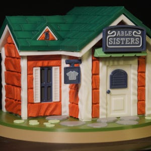 Puede incluir: Una maqueta en miniatura de la tienda Sable Sisters de Animal Crossing. El edificio presenta una fachada naranja, un tejado verde y un letrero que dice "Sable Sisters". Un pequeño letrero con un gráfico de camiseta cuelga afuera.