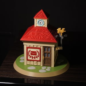 Puede incluir: Una maqueta en miniatura de un edificio con tejado rojo y torre del reloj. El edificio tiene una fachada beige con un símbolo de correo y una puerta de madera. Se asienta sobre una base verde con detalles de piedra blanca.