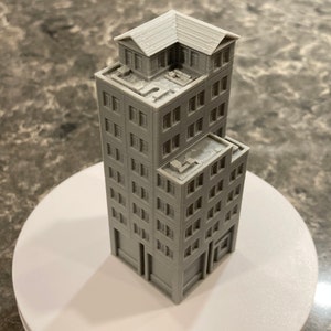 May include: A gray 3D printed model of a tall building with multiple floors and a rooftop addition. The building has a simple design with rectangular windows and a flat roof.