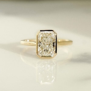 May include: A gold engagement ring with a square-shaped diamond set in a bezel setting.
