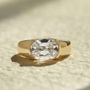 May include: A gold ring with a large, rectangular-cut diamond. The diamond is set in a bezel setting, and the band is a simple, polished gold. The ring is displayed on a textured, light-colored surface.