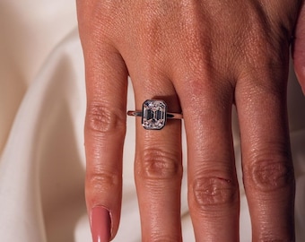 May include: A silver ring featuring an emerald-cut diamond. The ring is set on a thin band and is worn on a finger. The diamond has a rectangular shape with a step-cut facet pattern. The ring is a piece of jewellery.