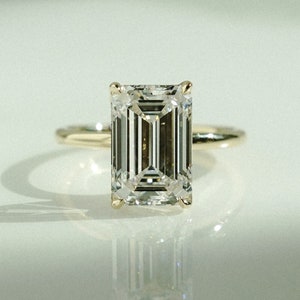 May include: A gold ring featuring a large, clear emerald-cut diamond. The diamond's facets catch the light, creating a brilliant display. The ring sits on a white surface, with its reflection clearly visible below.