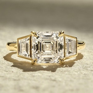 May include: A gold ring featuring a large, square-cut diamond center stone flanked by two smaller, rectangular diamonds. The ring's band and setting are a warm gold color, creating a classic and elegant look. The diamonds sparkle, reflecting light.