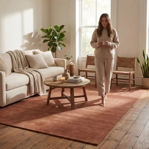 May include: A living room with a rust-coloured rectangular rug. A beige sofa with cushions and a throw is next to a round wooden coffee table. Two woven chairs and a woman holding a mug are also visible in the room.