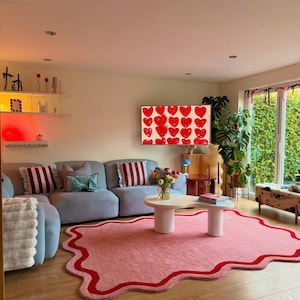 Könnte beinhalten: Ein Wohnzimmer mit hellblauem Sofa, rosa Teppich mit rotem Rand und weißem Couchtisch. Ein Fernseher zeigt rote Herzen. Dekorative Elemente sind Kissen, Pflanzen und Wandregale.