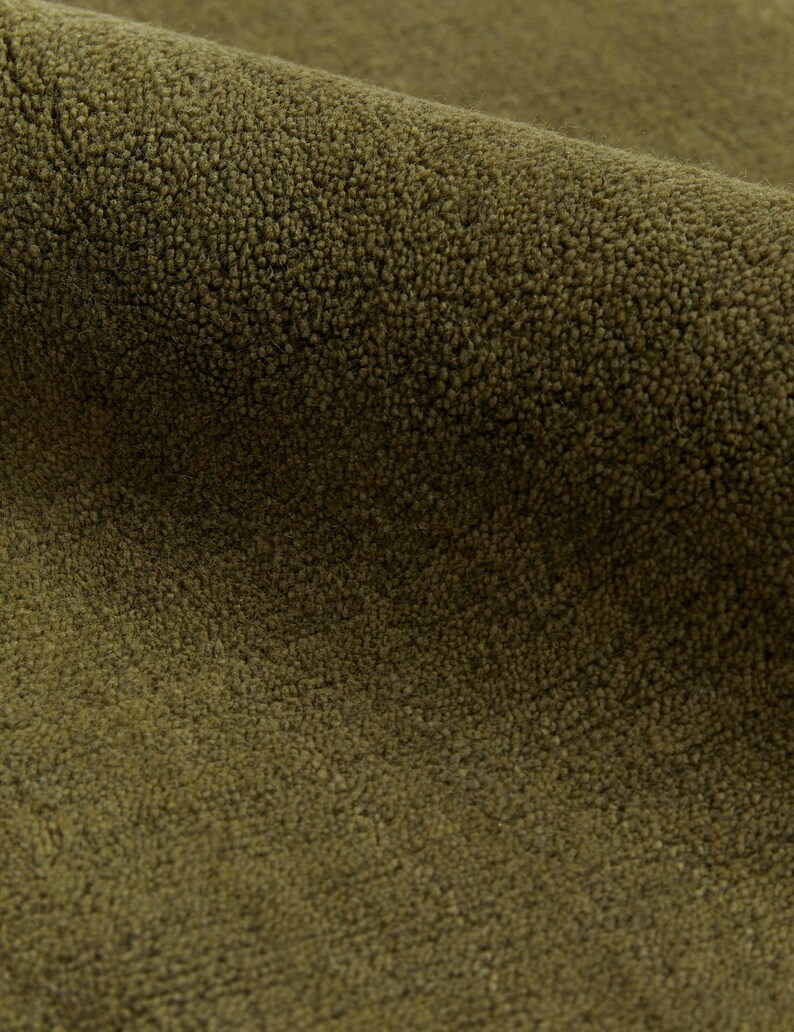 May include: Close-up of a section of olive green fabric with a textured, looped pile. The fabric is folded, revealing the depth and density of the material. The color is a muted, earthy tone, suggesting a natural fiber.