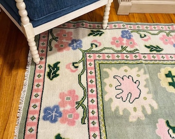 May include: A rectangular rug with a floral pattern in pink, blue, and green. The rug has a white background with a border of pink squares and green vines. The rug is placed on a wooden floor.