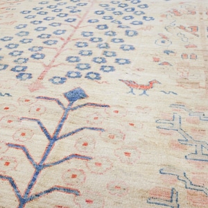 May include: A cream-colored rug with a repeating pattern of stylized trees in blue and pink. The trees have a diamond-shaped top and branches that extend out to the sides. The rug has a subtle texture and a worn look.