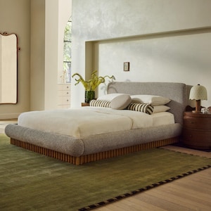 May include: A modern bedroom scene featuring a large bed with a gray upholstered headboard and base. The bed is made with white linens and decorative pillows. A green rug with a dark border sits on the wooden floor. A wooden nightstand with a lamp is next to the bed.