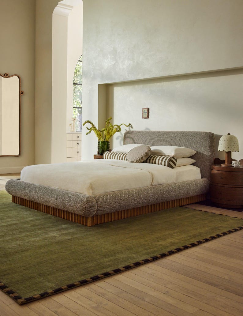 May include: A modern bedroom scene featuring a large bed with a gray upholstered headboard and base. The bed is made with white linens and decorative pillows. A green rug with a dark border sits on the wooden floor. A wooden nightstand with a lamp is next to the bed.
