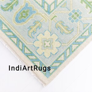 May include: Close-up of a corner of a light blue and green patterned rug with a white fringe. The rug has a geometric design with floral motifs.