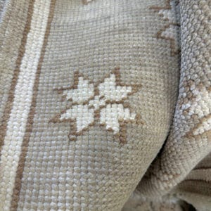 Pode incluir: Close-up de um tapete bege com um padrão floral repetido em branco sujo e castanho. O tapete tem uma superfície texturizada e uma borda listrada. O tapete está dobrado, revelando a parte inferior.