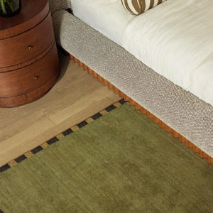 May include: A bedroom scene featuring a bed with a patterned headboard and a green rug with a black and gold border. A wooden nightstand with drawers is visible, along with a striped bolster pillow.